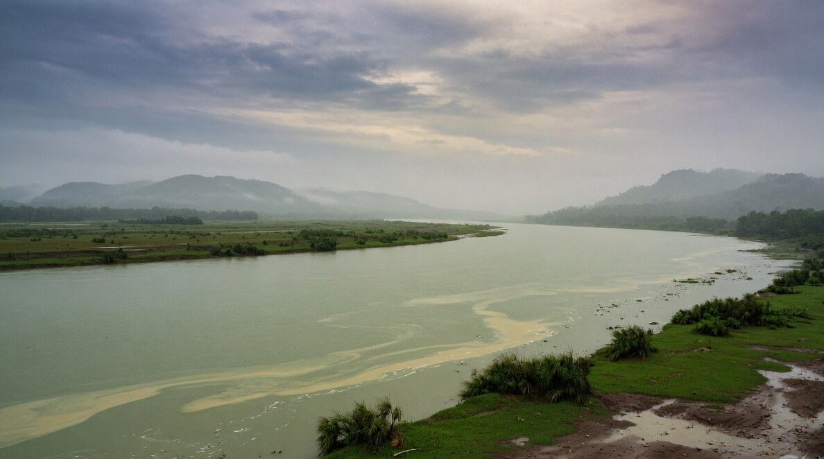 Wide river landscape in Assam under monsoon cloud, looking across silt-green water toward misted hills